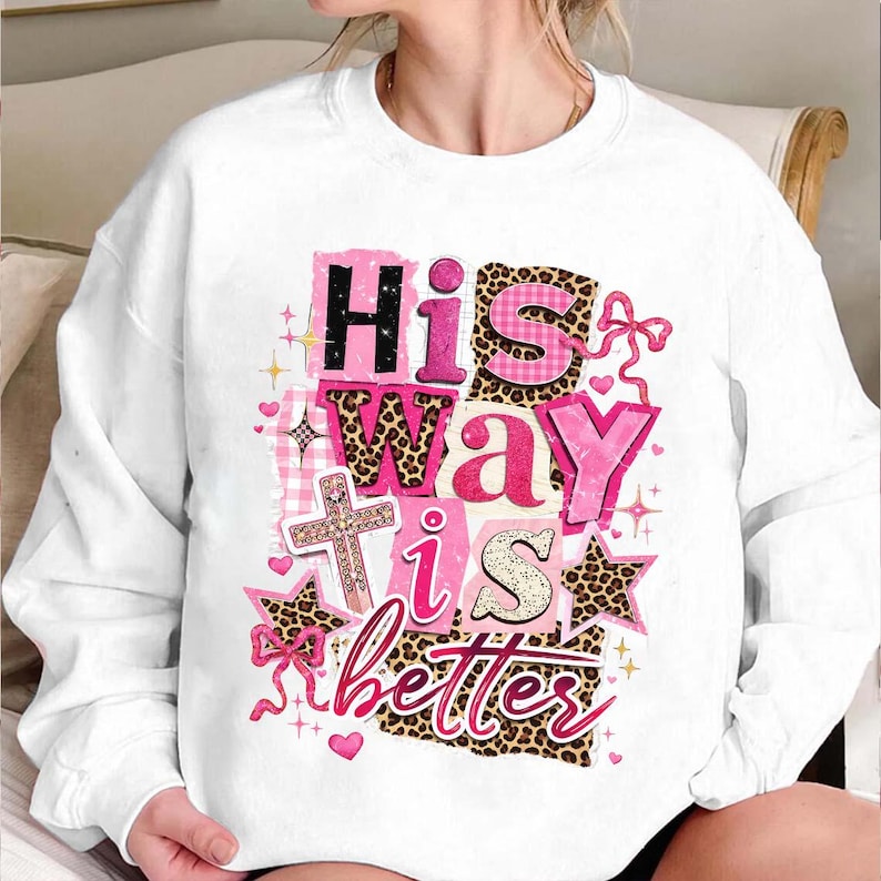 May include: White sweatshirt with the text "His Way is Better" in pink, black, and leopard print. The design includes a cross, stars, and bows. The sweatshirt is a casual, comfortable style.