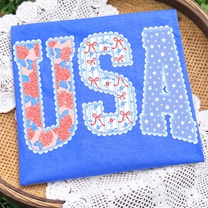 Puede incluir: Una camiseta azul con la palabra "USA" en letras decorativas. La "U" está llena de patrones florales rojos y rosas, la "S" tiene rayas azules y lazos rojos, y la "A" está llena de estrellas blancas.