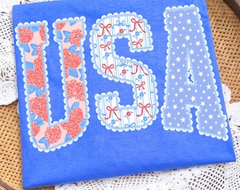 May include: A blue t-shirt with the word "USA" in decorative letters. The "U" is filled with red and pink floral patterns, the "S" has blue stripes and red bows, and the "A" is filled with white stars.