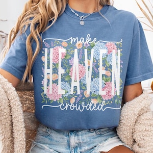 May include: A blue t-shirt with the words "make HEAVEN crowded" in white, surrounded by a floral design of pink, blue, and peach flowers. The shirt is a solid blue color.