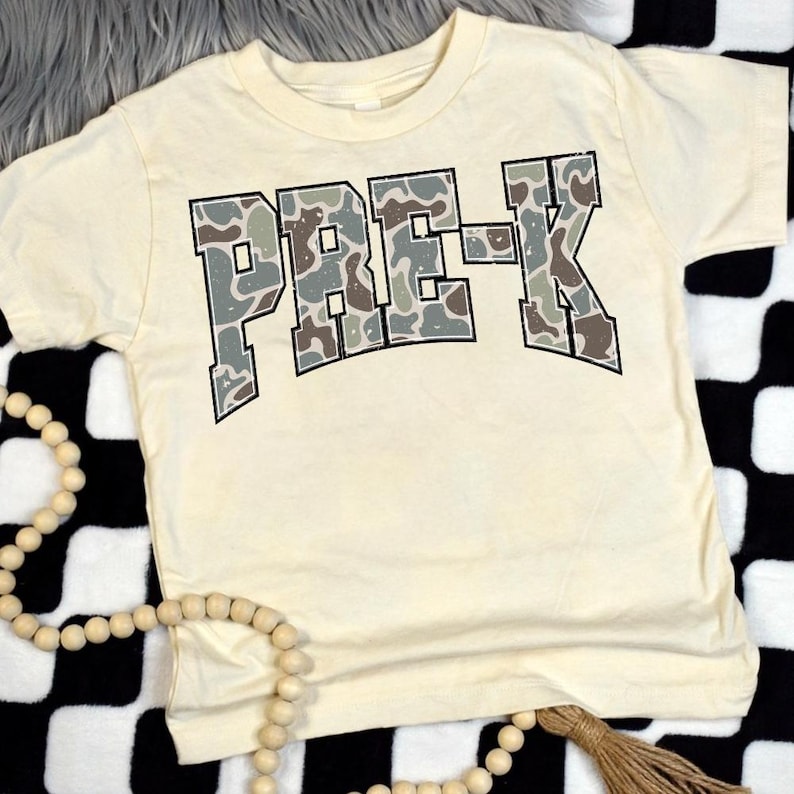 May include: Cream-colored short-sleeved t-shirt with the text "PRE-K" in a camouflage pattern. The letters are outlined in black. The shirt is laying on a black and white checkered surface.