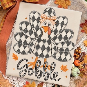 May include: A cream-colored fabric featuring a graphic of a turkey with black and white buffalo plaid feathers, an orange beak, and a small orange and white gingham bow on its head. The word GOBBLE is printed in black script below the turkey.