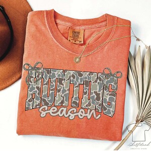 May include: Orange Comfort Colors t-shirt with the words "HUNTING season" in a camouflage pattern. The shirt has a crew neck and short sleeves. A gold necklace with a pendant is draped on the shirt.