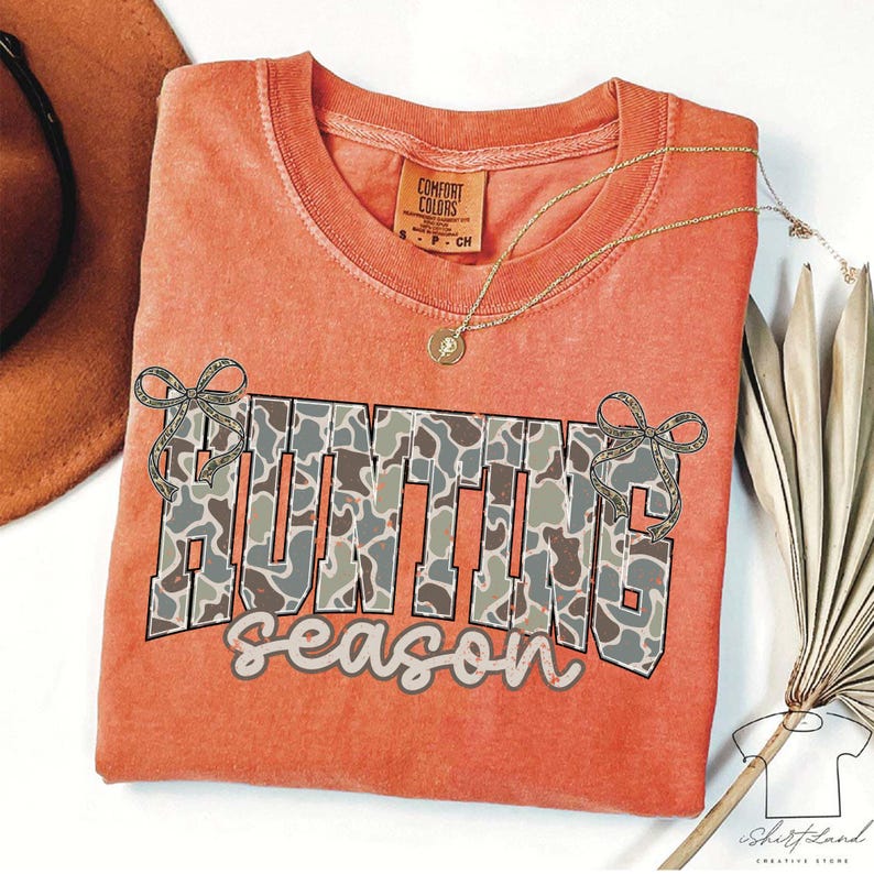 May include: Orange Comfort Colors t-shirt with the words "HUNTING season" in a camouflage pattern. The shirt has a crew neck and short sleeves. A gold necklace with a pendant is draped on the shirt.