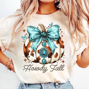 May include: A white t-shirt with a brown and white cow print pumpkin with a turquoise bow and rhinestones. The text "Howdy Fall" is written in brown with a rope design around it.