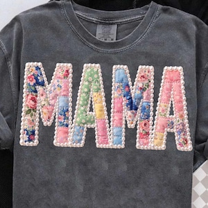 以下が含まれることがあります： 前面に「MAMA」の文字がアップリケされた濃いグレーのスウェットシャツ。文字は様々な花柄と色合いのパッチワーク生地で作られ、小さな白いビーズで縁取られています。Comfort Colorsのタグが見えます。