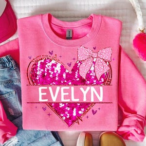 May include: Pink sweatshirt with a sequined heart and a bow. The heart has the name "EVELYN" written on it.