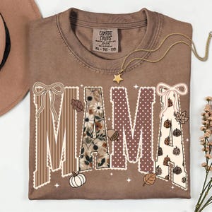 Peut inclure: Un t-shirt marron &agrave; manches courtes avec le mot MAMA imprim&eacute; en grandes lettres. Chaque lettre pr&eacute;sente un motif diff&eacute;rent, notamment des rayures, des fleurs, des pois et des citrouilles l&eacute;opard, agr&eacute;ment&eacute; de n&oelig;uds, feuilles et &eacute;toiles.
