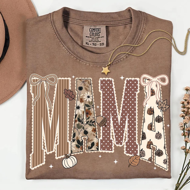 Peut inclure: Un t-shirt marron &agrave; manches courtes avec le mot MAMA imprim&eacute; en grandes lettres. Chaque lettre pr&eacute;sente un motif diff&eacute;rent, notamment des rayures, des fleurs, des pois et des citrouilles l&eacute;opard, agr&eacute;ment&eacute; de n&oelig;uds, feuilles et &eacute;toiles.