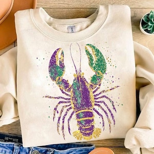May include: A cream-colored sweatshirt with a purple, green, and gold glitter lobster design. The lobster is in a standing position with its claws raised.
