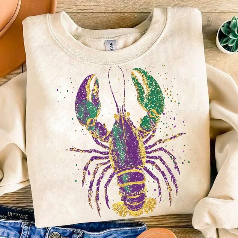 May include: A cream-colored sweatshirt with a purple, green, and gold glitter lobster design. The lobster is in a standing position with its claws raised.