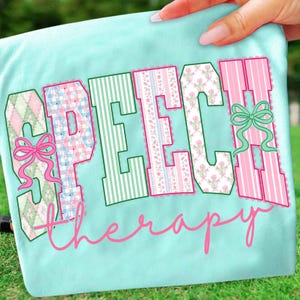 Puede incluir: Una camiseta azul claro con las palabras "SPEECH THERAPY" en una fuente decorativa. Las letras est&aacute;n rellenas con varios patrones, incluyendo rayas, estampados florales y cuadros. Lazos rosas y verdes acent&uacute;an el dise&ntilde;o.