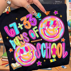 Może przedstawiać: Czarna bluza z kolorową grafiką. Projekt zawiera napis "67 + 33 DAYS OF SCHOOL" oraz dwie uśmiechnięte buzie z błyskawicami. Dodatkowo gwiazdki, oł&oacute;wki i globus.