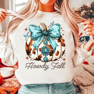 May include: White sweatshirt with a brown and white cow print pumpkin with a blue bow and rhinestones. The text "Howdy Fall" is written below the pumpkin.