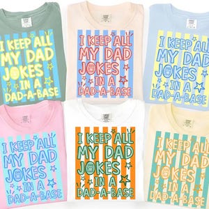 May include: Six folded t-shirts in various colors, including green, pink, and white, each featuring the text "I KEEP ALL MY DAD JOKES IN A DAD-A-BASE" in a colorful, retro design. The shirts are made of soft material.