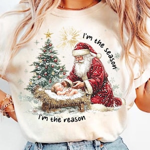 Puede incluir: Camiseta color crema con un gráfico navideño. El diseño incluye a Papá Noel, un bebé en un pesebre y un árbol de Navidad decorado. El texto dice "I'm the season, I'm the reason."