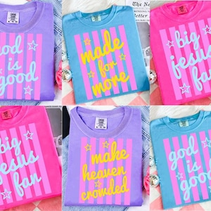 May include: A collection of colorful t-shirts in shades of purple, blue, and pink. Each shirt features a unique graphic design with text and stars. The text includes phrases like "God is Good" and "Big Jesus Fan."