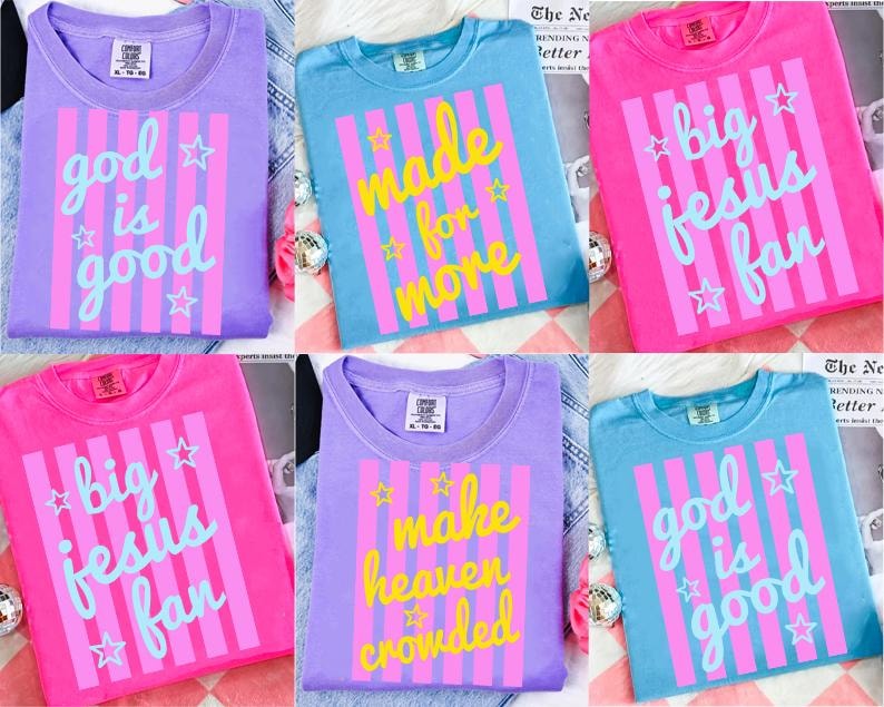 May include: A collection of colorful t-shirts in shades of purple, blue, and pink. Each shirt features a unique graphic design with text and stars. The text includes phrases like "God is Good" and "Big Jesus Fan."