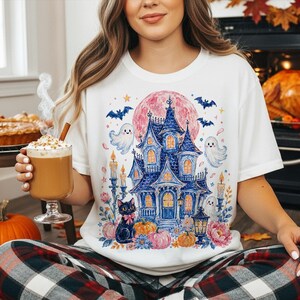 Puede incluir: Una camiseta blanca de manga corta con un gr&aacute;fico colorido de una casa encantada azul, un gato negro con un lazo rosa, fantasmas, murci&eacute;lagos, calabazas y una gran luna rosa, rodeados de velas encendidas y flores.