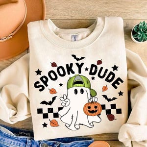 May include: A cream colored sweatshirt with a graphic of a ghost wearing a green baseball cap and holding a jack-o-lantern. The text "Spooky Dude" is printed in black above the ghost.