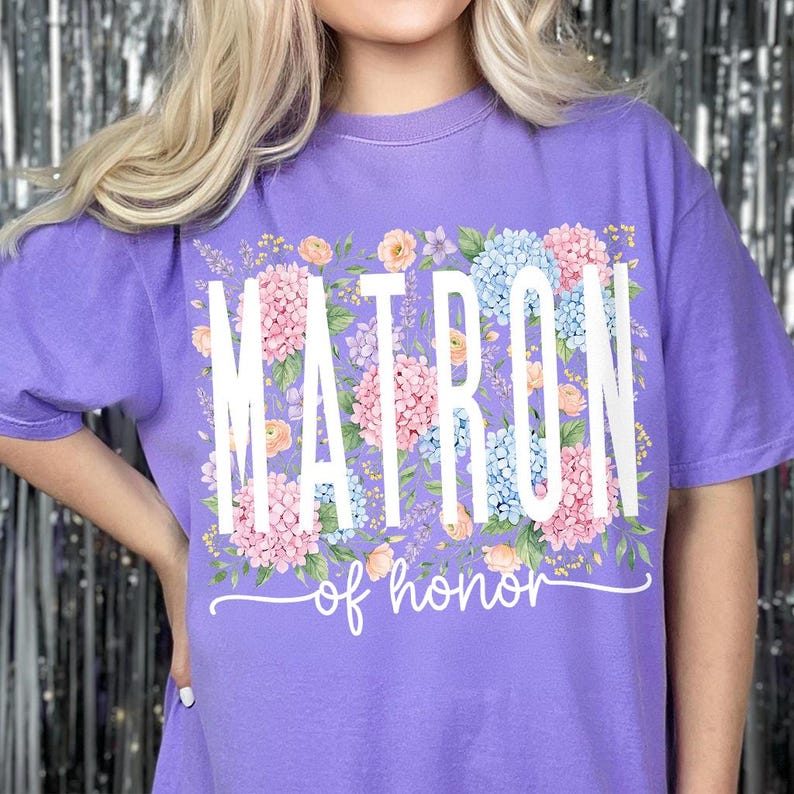 May include: A purple t-shirt with the word "MATRON" in large white letters, surrounded by a floral design of pink, blue, and peach flowers. Below the word "MATRON" is the phrase "of honor" in a cursive font.