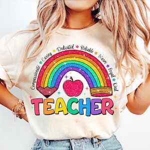 Könnte beinhalten: Ein weißes T-Shirt mit einem Regenbogen und einem roten Apfel mit Glitzer. Der Regenbogen ist von den Wörtern "Compassionate", "Caring", "Dedicated", "Reliable", "Warm", "Loyal" und "Kind" umgeben. Die Wörter "TEACHER" befinden sich unter dem Regenbogen.