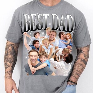 May include: Gray t-shirt with the text "BEST DAD" in a silver gradient font. The shirt features a collage of photos of fathers with their children. The overall theme is family and fatherhood.