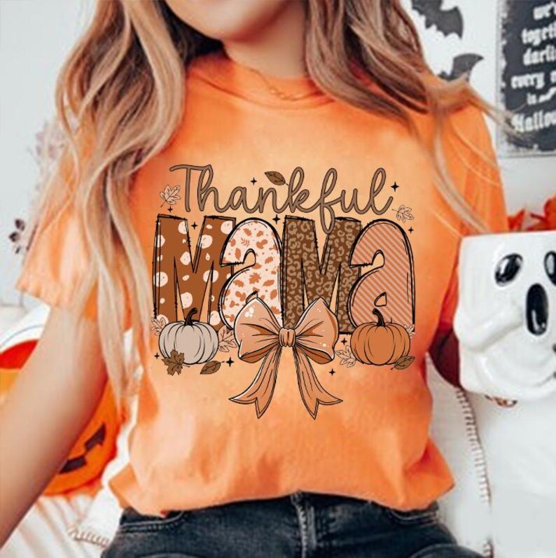 Op de afbeelding: Oranje T-shirt met de tekst "Thankful Mama" in bruine letters met een patroon. De letters zijn versierd met pompoenen, bladeren en een bruine strik.