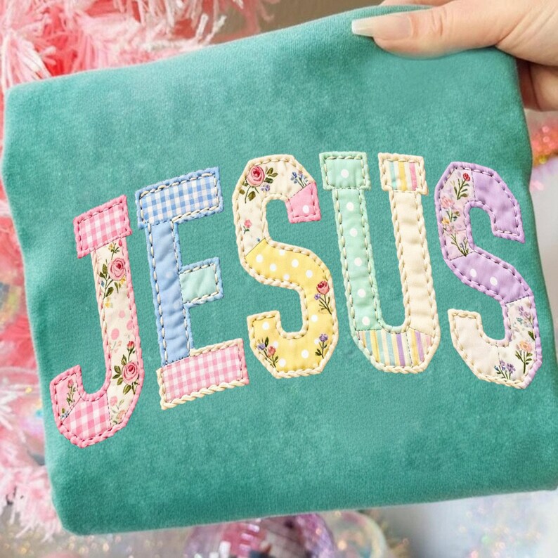 Pode incluir: Tecido verde-azulado com a palavra "JESUS" aplicada em letras de patchwork. Cada letra apresenta um padr&atilde;o diferente, incluindo estampas florais, bolinhas e xadrez, com bordas onduladas.
