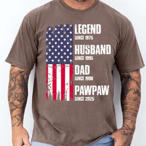 K&ouml;nnte beinhalten: Braunes T-Shirt mit einem verblassten Design der amerikanischen Flagge. Das Shirt zeigt die Worte "LEGEND SINCE 1975, HUSBAND SINCE 1995, DAD SINCE 1998, PAWPAW SINCE 2025" in wei&szlig;en Blockbuchstaben.