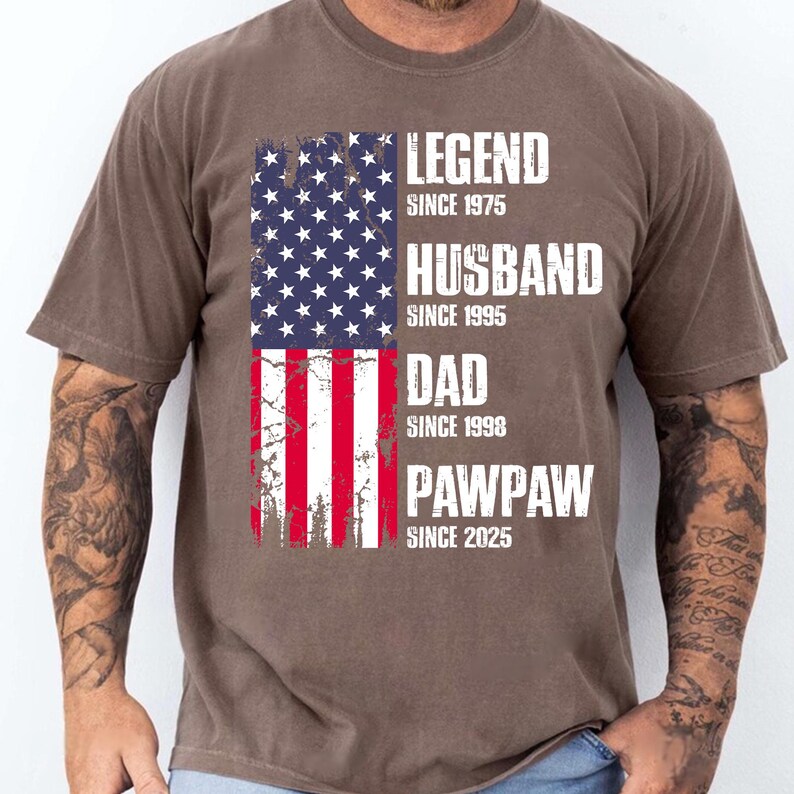 K&ouml;nnte beinhalten: Braunes T-Shirt mit einem verblassten Design der amerikanischen Flagge. Das Shirt zeigt die Worte "LEGEND SINCE 1975, HUSBAND SINCE 1995, DAD SINCE 1998, PAWPAW SINCE 2025" in wei&szlig;en Blockbuchstaben.