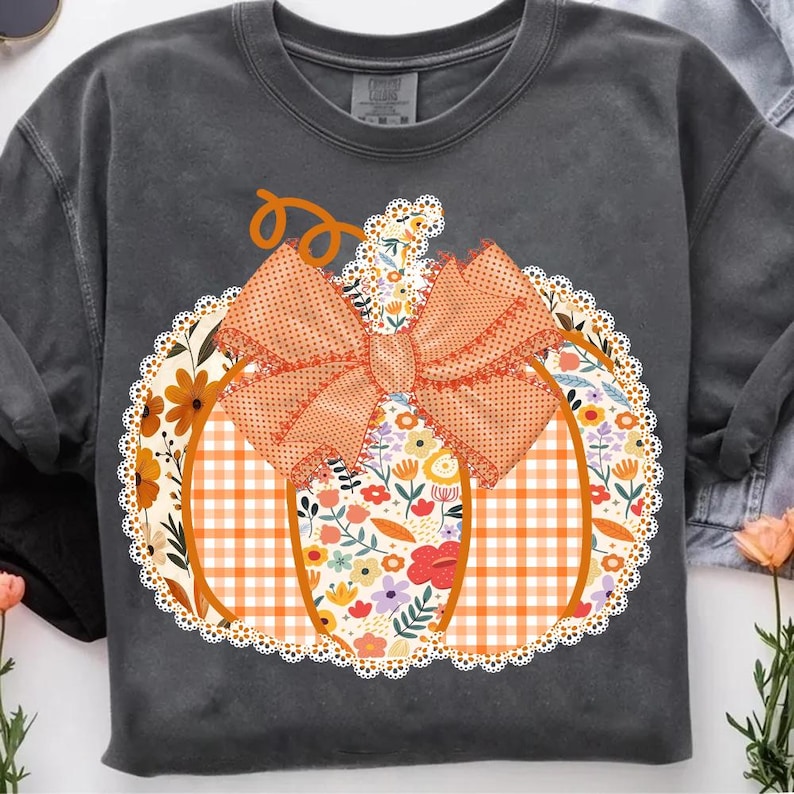 K&ouml;nnte beinhalten: Dunkelgraues Sweatshirt mit einem K&uuml;rbisdesign. Der K&uuml;rbis hat eine orangefarbene Schleife und ein Blumen- und Karomuster. Das Design ist von einem wei&szlig;en Spitzensaum umgeben, ideal f&uuml;r den Herbst.