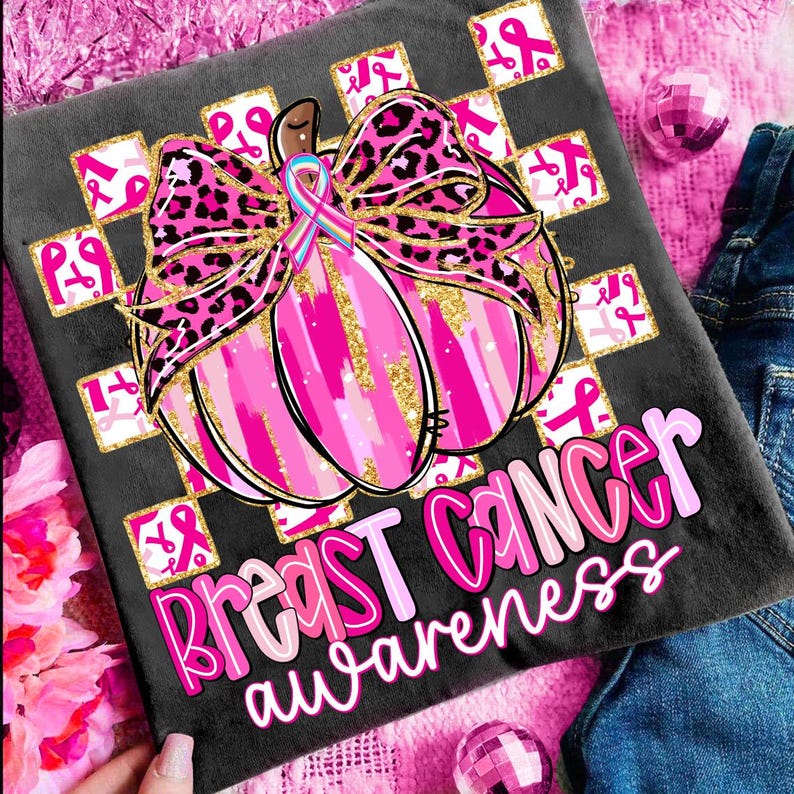 May include: A dark gray t-shirt with a pink and gold pumpkin design, a leopard print bow, and the words "Breast Cancer Awareness" in pink. The design includes pink ribbons and a blue and pink ribbon.