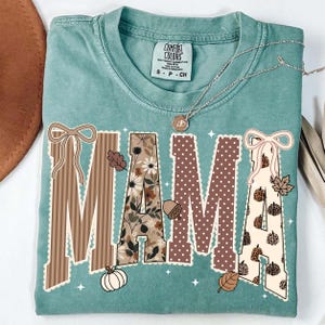 Peut inclure: Un t-shirt &agrave; col rond bleu sarcelle avec le mot MAMA en grandes lettres majuscules. Chaque lettre pr&eacute;sente un motif unique, notamment des rayures brunes, des fleurs, des pois bruns et des citrouilles &agrave; imprim&eacute; l&eacute;opard, avec des n&oelig;uds.