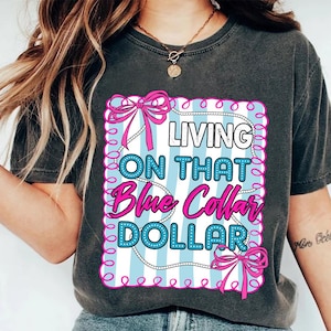 May include: Dark gray t-shirt with the text "LIVING ON THAT Blue Collar DOLLAR" in a pink and blue design. The design includes bows and a pearl necklace-like border. The shirt is a casual wear item.