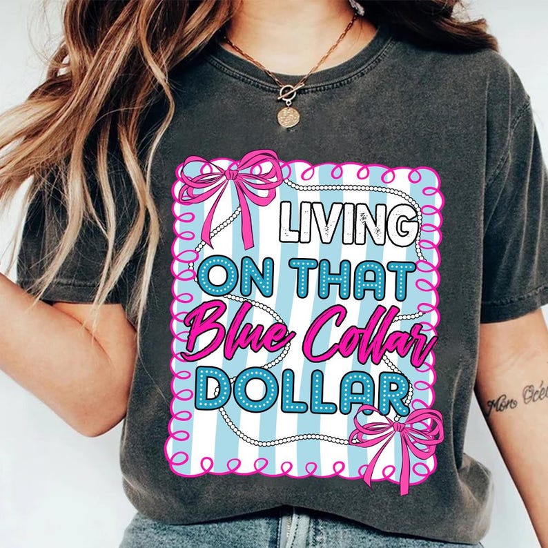 May include: Dark gray t-shirt with the text "LIVING ON THAT Blue Collar DOLLAR" in a pink and blue design. The design includes bows and a pearl necklace-like border. The shirt is a casual wear item.