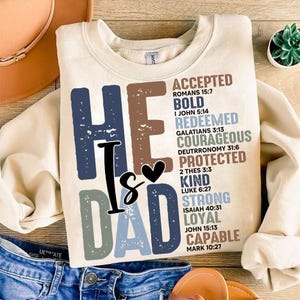May include: Cream-colored sweatshirt with the text "HE IS DAD" in blue, brown, and gray. The words "ACCEPTED, BOLD, REDEEMED, COURAGEOUS, PROTECTED, KIND, STRONG, LOYAL, CAPABLE" are also printed on the shirt.