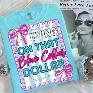 May include: A light blue Comfort Colors t-shirt with the phrase "LIVING ON THAT Blue Collar DOLLAR" in a decorative design. The design includes pink bows, white stripes, and a pearl-like border. The shirt is folded on a white surface.