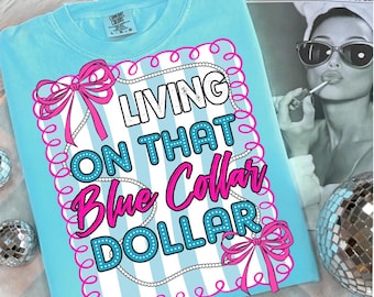 Puede incluir: Una camiseta Comfort Colors azul claro con la frase "LIVING ON THAT Blue Collar DOLLAR" en un diseño decorativo. El diseño incluye lazos rosas, rayas blancas y un borde de perlas. La camiseta está doblada sobre una superficie blanca.