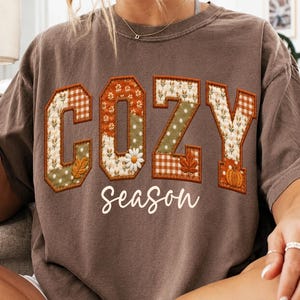 May include: A brown short-sleeve t-shirt featuring the word COZY in large, block-style letters filled with various autumn-themed fabric patterns, including plaid, polka dots, and floral prints. Below, the word season is printed in white script.