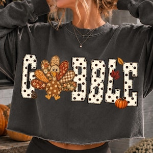 May include: A charcoal gray long-sleeve cropped sweatshirt featuring the word GOBBLE in large white letters with black polka dots. The letter O is replaced by a turkey with patterned feathers. A small orange pumpkin and autumn leaves are included.