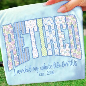 May include: Light blue t-shirt with the word "RETIRED" in large, decorative letters. The letters feature floral and striped patterns in shades of blue, green, and silver. Below the word, the text reads "I worked my whole life for this" and "Est. 2026".