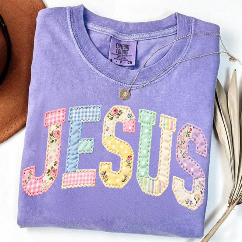 Pode incluir: Uma camiseta Comfort Colors lavanda com a palavra "JESUS" aplicada em tecido patchwork. As letras apresentam padr&otilde;es florais, xadrez e de bolinhas. Um colar de prata com um pingente repousa na camisa.