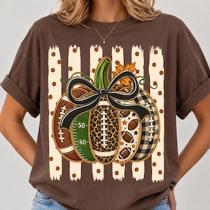May include: A brown short-sleeve t-shirt featuring a graphic of a pumpkin composed of football-themed segments, including a green field section with 40 and 50 yard line markers, a leopard print section, and a black and white buffalo plaid section.