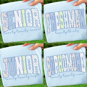 May include: Four light blue t-shirts featuring collegiate-style lettering in floral and gingham patterns. The shirts display the words SENIOR twenty twenty-seven, FRESHMAN twenty thirty, JUNIOR twenty twenty-eight, and SOPHOMORE twenty twenty-nine.