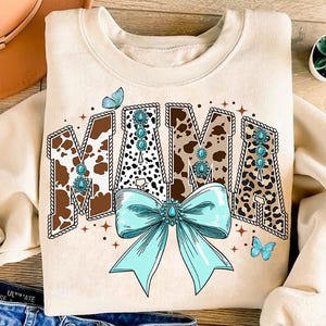 Op de afbeelding: Een beige sweatshirt met een grafische print van het woord "MAMA" in bruine, zwarte en witte dierenprintletters. De letters zijn omrand met bruin touw en versierd met turquoise edelstenen. Een turquoise strik met een grote turquoise edelsteen in het midden bevindt zich onder het woord "MAMA". Twee blauwe vlinders bevinden zich aan weerszijden van de grafiek.