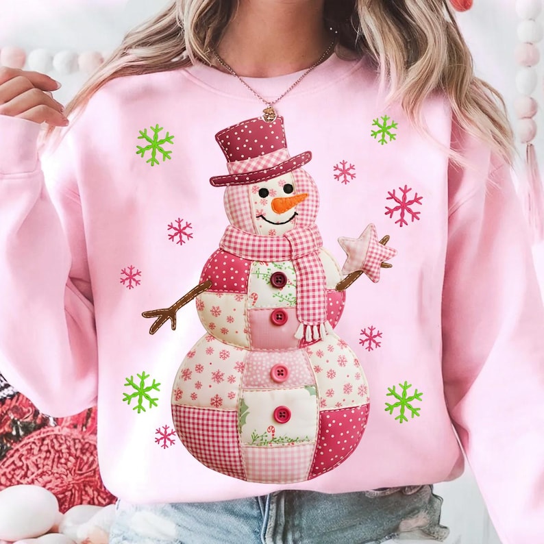 May include: Pale pink sweatshirt with a patchwork snowman design. The snowman has a carrot nose, a plaid scarf, and a star-shaped hand. Green and pink snowflakes are scattered around the snowman. The snowman is wearing a red polka dot top hat.