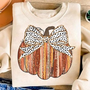 May include: A cream-colored sweatshirt with a graphic of a pumpkin with a black and white polka dot bow. The pumpkin is decorated with orange, brown, and gold stripes.