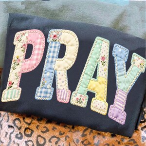 Könnte beinhalten: Dunkelblauer Stoff mit dem Wort "PRAY" in bunten Patchwork-Buchstaben appliziert. Jeder Buchstabe hat eine andere Pastellfarbe und ein anderes Muster, darunter Blumen, Vichy-Karo und Punkte. Die Buchstaben sind mit einer weißen Naht umrandet.