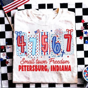 May include: Cream-colored t-shirt with a patriotic design featuring the numbers 4, 7, 5, 6, and 7 in red and blue. The shirt includes the text "Small town Freedom PETERSBURG, INDIANA" in red. An American flag is visible.